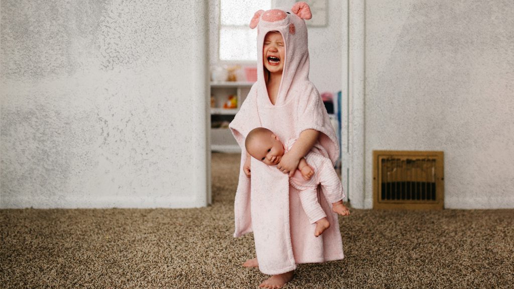 A child crying during a tantrum. They are wearing a pale pink bathrobe and holding a doll.